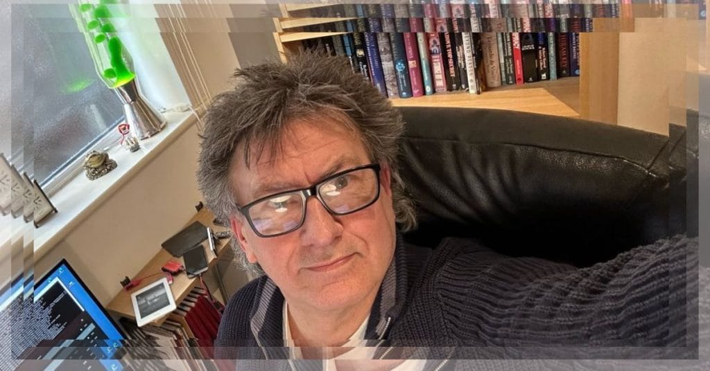 A man sitting at a desk with books in the background , taking a selfie of himself